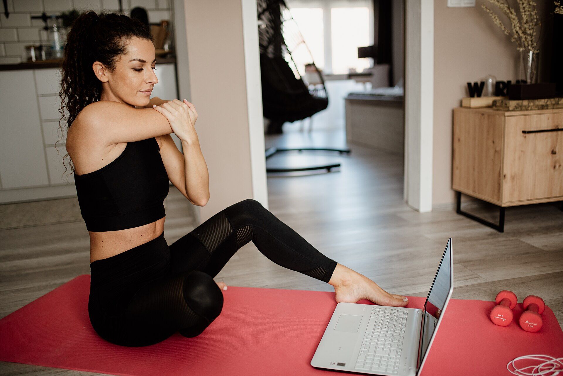 A woman sitting on an exercise mat at home, following a physiotherapy session on a laptop.