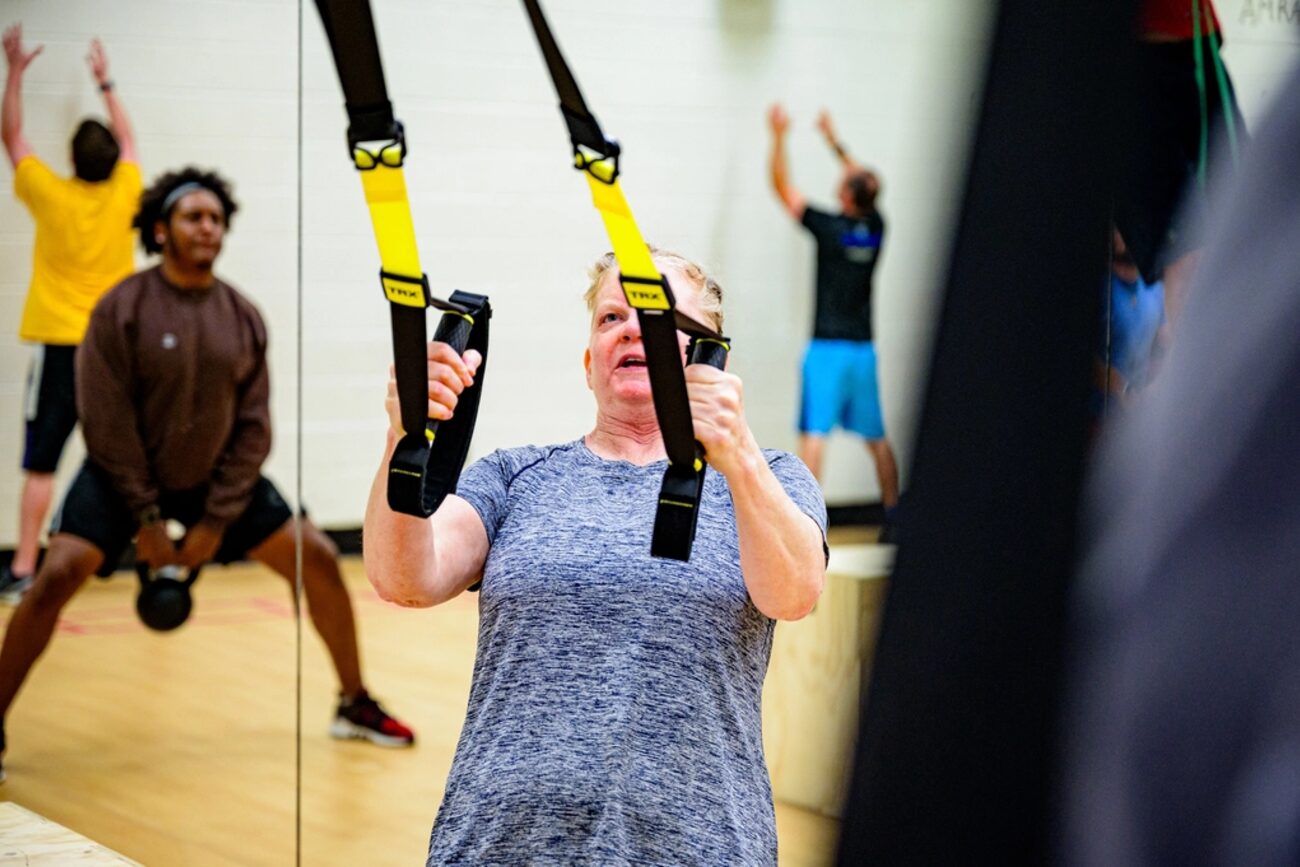 An older adult training with suspension straps in a gym under supervision.